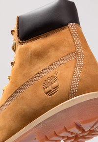 Bottine en suède tan avec un col noir, des coutures blanches, un logo d'arbre bien visible et une semelle en caoutchouc robuste dotée d'un motif de crampons.