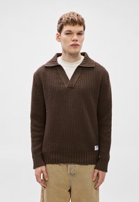 Barna kötött pulóver texturált mintával, V-nyakkal és széles bordázott mandzsettákkal. Csíkos gallérral és kis márkajelzéssel az alsó szélén.