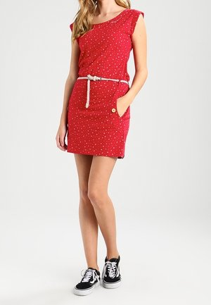 Vrouw in een mouwloze rode mini-jurk met een wit bloemmotief, een witte riem en zwarte sneakers, staande met één hand in de zak.