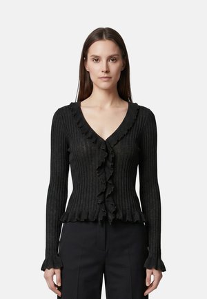 Donna con capelli castani lisci indossa un cardigan nero a coste aderente con bordi arricciati e pantaloni neri, in piedi davanti a uno sfondo semplice.