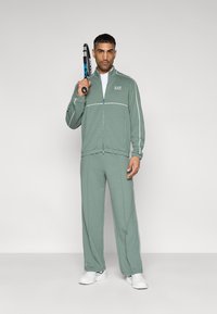 Πράσινη αθλητική φόρμα με λευκές ρίγες, φτιαγμένη από απαλό ύφασμα. Μπουφάν με φερμουάρ και λογότυπο, σετ με φαρδιά παντελόνια και λευκά αθλητικά παπούτσια.