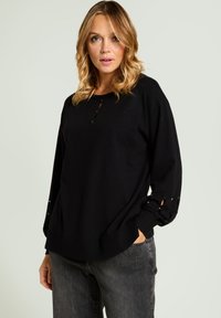 Suéter de punto negro con cuello redondeado, detalle de botones en el pecho y cortes decorativos en los puños. Combinado con vaqueros grises.