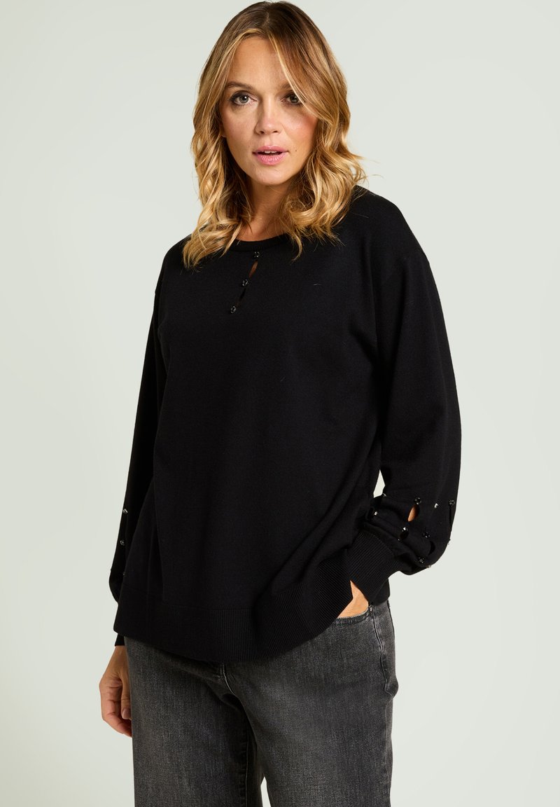 Suéter de punto negro con cuello redondeado, detalle de botones en el pecho y cortes decorativos en los puños. Combinado con vaqueros grises.