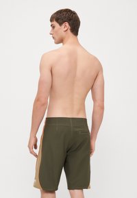 Grüne Badeshorts mit orangen Seitenstreifen, knielang, mit einer Gesäßtasche und glatter Textur. Minimalistisches Design, ohne sichtbare Markenkennung.
