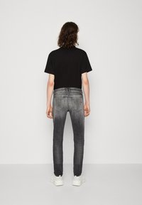 Persona con cabello rizado que lleva una camiseta negra, jeans grises desgastados y zapatillas blancas, de pie frente a una pared gris claro liso.