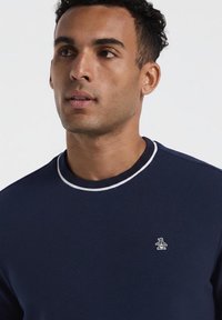 Homme aux cheveux courts et bouclés portant un pull ras du cou bleu marine avec un bord blanc et un petit logo de pingouin brodé sur la poitrine.