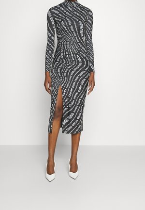 Kvinde iført en sort og hvid langærmet midikjole med "HUGO" tekstmønster og frontslids op til låret, kombineret med hvide spidse hæle.