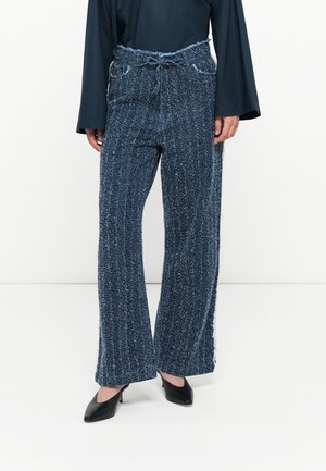 Tweed trousers in dark blue with a textured finish, wide legs, and a frayed waistband. Paired with a long-sleeve top and black heels.