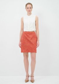 Blouse blanche sans manches avec un devant à boutons, associée à une jupe texturée orange-rouge arrivant au genou. Des talons marron viennent compléter le look.