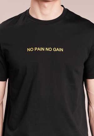 Homme portant un t-shirt noir avec le texte jaune "NO PAIN NO GAIN" centré sur la poitrine, recadré au niveau du cou et au milieu des bras.