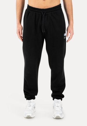 Uomo che indossa pantaloni da jogging neri con polsini elastici e sneakers bianche, in piedi contro uno sfondo bianco uniforme.