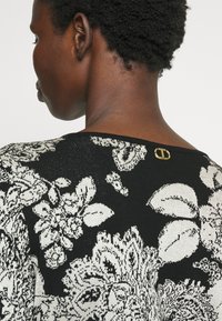 Persoon met kort, krullend haar draagt een zwarte top met een wit bloemenpatroon en een kleine gouden sluiting op de achterkant van de halslijn.