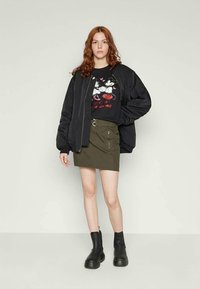 Ung kvinna med rött hår iklädd en svart oversized jacka, t-shirt med Mickey och Minnie Mouse grafik, olivgrön kjol och svarta ankelstövlar.