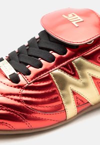 Κόκκινο μεταλλικό αθλητικό παπούτσι με μαύρα κορδόνια, χρυσό λογότυπο "M" στο πλάι και κόκκινη γλώσσα με χρυσό έμβλημα "ML".