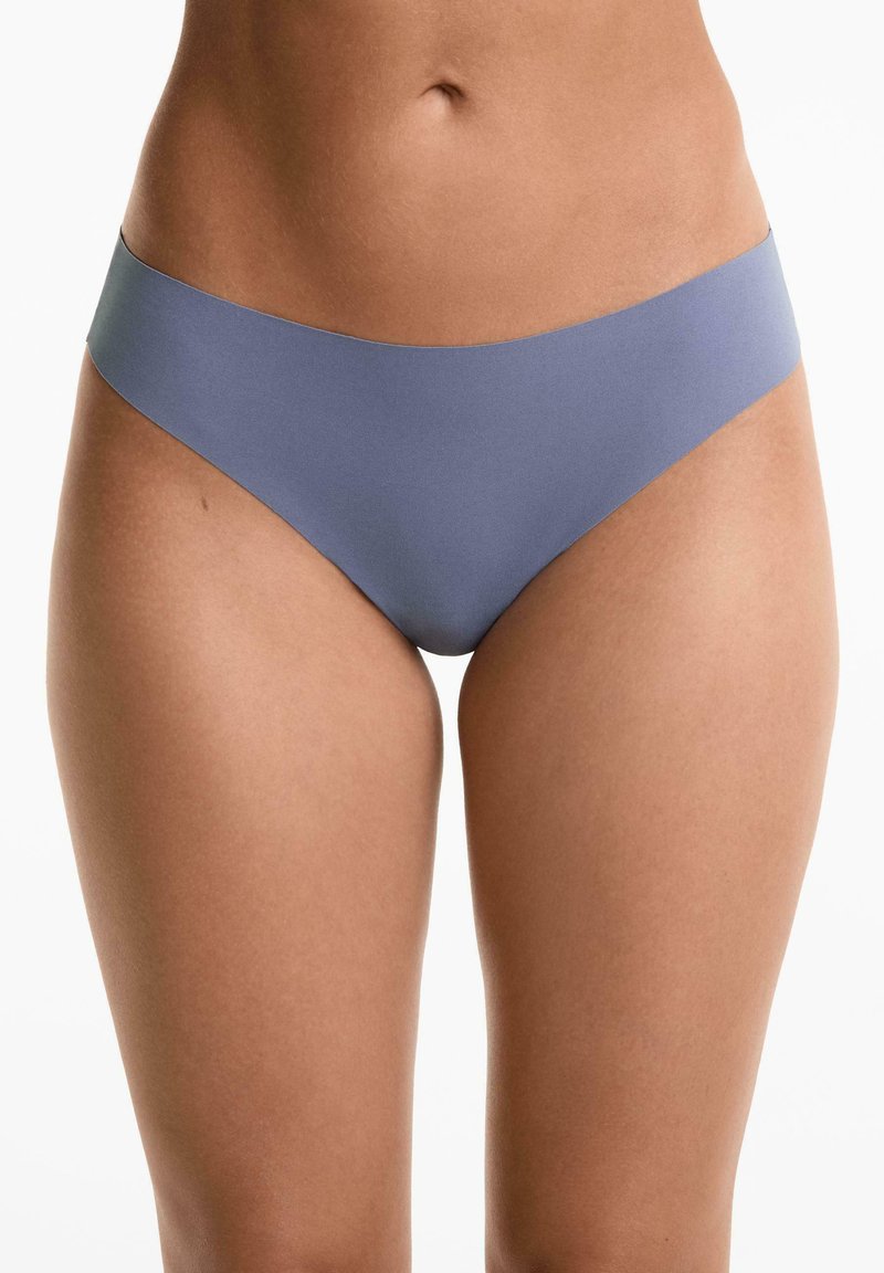 Vrouw in naadloze lichtblauwe bikini-achtige onderbroek, die taille, heupen en bovenbenen toont tegen een witte achtergrond.