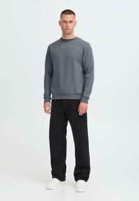 Man staat in een grijs sweatshirt, losse zwarte broek en witte sneakers tegen een effen lichte achtergrond.