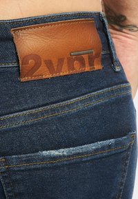 Blå denimjeans med gule sømmer, en tanfarget skinnmerkelapp med teksten "2YDR" på midjebåndet, og en tatovert hånd hvilende nær lommen.