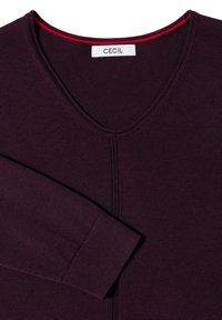 Pull violet foncé en tricot avec col en V et manches longues repliées sur la poitrine, montrant une étiquette blanche avec le nom de la marque Cecil.