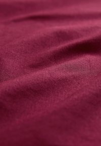 Głęboki burgundowy materiał o gładkiej fakturze, z delikatnymi zmarszczkami i drobnym wzorem splotu, który tworzy głębię i wymiary.
