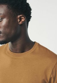 Brun bomulds t-shirt med rund halsudskæring, der har en tekstureret overflade og korte ærmer, præsenteret på en tekstureret neutral baggrund.