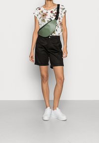 Grön konstläder crossbody-väska med svart rem, blommig vit topp, svarta shorts och vita sneakers. Enkel design med rundad form.