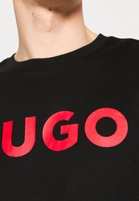 Schwarzes T-Shirt mit weitem Ausschnitt, das in kräftigen, leuchtend roten Buchstaben "UGO" über die Brust gedruckt hat. Weicher Baumwollstoff, minimalistisches Design.