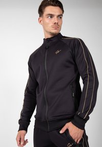 Zwarte sportieve zipjack met gouden strepen, slim-fit ontwerp, hoge kraag en zijzakken met ritssluiting. Voorzien van merklogo op de borst.