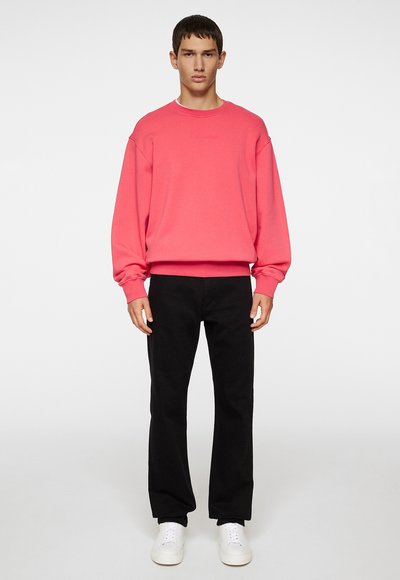 Felpa rosa con polsini e orlo a coste, abbinata a pantaloni neri e sneakers bianche. Il modello si trova di fronte a uno sfondo bianco.