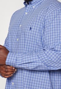 Niebiesko-biała koszula w kratę na guziki z długimi rękawami, z klasycznym kołnierzykiem i logo na piersi. Materiał bawełniany.