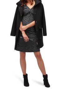 Zwarte overjas met capuchon, gedragen over een gemarmerde grijze jurk. De jurk heeft een verzamelde halslijn, gecombineerd met een zwarte clutch en enkelhoge laarzen.