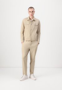Bēšas krāsas pogājama jaka ar diviem krūšu kabatām, saskaņota ar atbilstošām šaurām biksēm. Baltas sporta kurpes noslēdz apģērbu. Minimalistisks dizains.