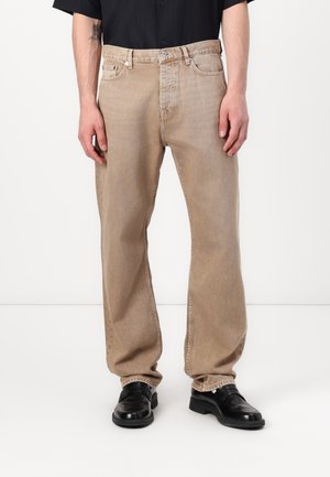 Homme portant un jean beige droit, une chemise noire et des chaussures en cuir noires, debout devant un fond blanc.