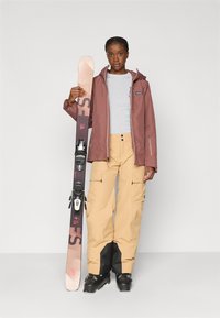 Conjunto de ski com uma jaqueta impermeável bordô, camisa cinza, calças cargo bege e botas de ski pretas, segurando um ski bicolor com fixações.