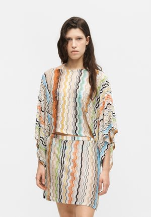 Femme aux longs cheveux foncés portant un haut ample à motif zigzag multicolore et une jupe assortie sur un fond blanc uni.