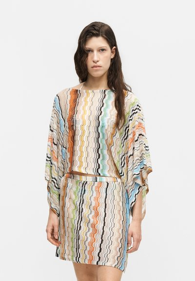 Femme aux longs cheveux foncés portant un haut ample à motif zigzag multicolore et une jupe assortie sur un fond blanc uni.