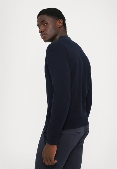 Homme portant un pull bleu marine et un pantalon foncé, tourné légèrement avec la tête tournée par-dessus son épaule sur un fond blanc uni.