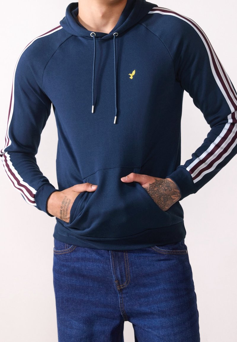 Sudadera azul marino con un bolsillo canguro, con acentos en franjas blancas y burdeos en las mangas y un pequeño logo amarillo en el pecho.