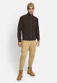 Brun strikket hættetrøje med lynlås og høj krave, beige cargo bukser og brune snørebåndsstøvler. Tilbehøret inkluderer en beige strikket hue og en sølv kæde.
