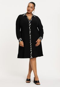 Zwarte jurk met lange mouwen, voorzien van een zebraprint kraag en manchetten. Het ontwerp omvat knopen langs de voorkant en een uitlopende rok.