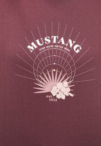 Fialová tkanina s bílým textem "MUSTANG pravý styl nikdy neumírá est. 1932" nad růžovými palmami a květinami s tečkovaným kruhovým vzorem.
