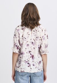 Blusa floreale bianca con motivi viola e neri, maniche a sbuffo, polsini arricciati e apertura centrale sul retro, abbinata a jeans in denim chiaro.