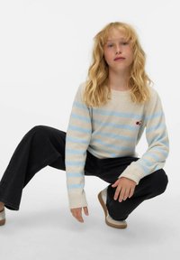 Pull crème avec des rayures bleues et un détail cerise ; associé à un jean noir à jambes larges et des chaussures de couleur claire. Texture douce, coupe décontractée.