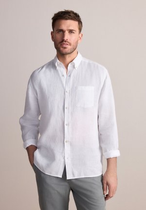 Camicia bianca di lino con design a bottoni, maniche lunghe arrotolate fino ai gomiti e una singola tasca sul petto. Abbinata a pantaloni grigi chiari.