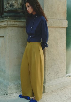 Femme aux cheveux bouclés portant une chemise bleu foncé boutonnée, un pantalon large couleur moutarde et des chaussures plates bleues, debout contre un pilier en pierre.