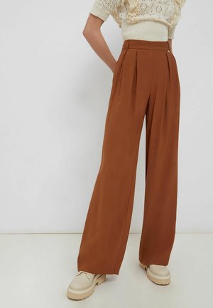 Kvinde iført højtaljede, vidde bukser i rust-brun farve kombineret med cremefarvede chunky-sålede sko og en struktureret cremefarvet sweater.