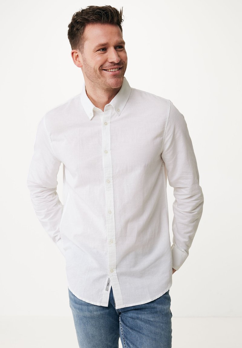 Camisa blanca de lino con botones, de manga larga, puños con un botón y dobladillo curvado. Lleva puesta con jeans azules, destacando un estilo casual.