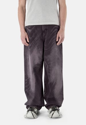 Osoba ubrana w luźne, ciemnofioletowe, sprane dżinsy oraz białe czarne trampki, stojąca na tle jednolitego jasnego tła.