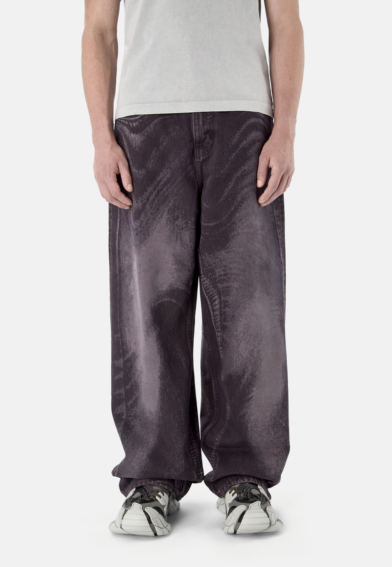 Person, die locker sitzende, dunkelviolette Jeans mit verwaschenem Look und weiße schwarze Sneaker trägt, vor einem schlichten hellen Hintergrund steht.