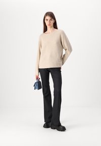 Šviesiai rudas megztinis su laisvu siluetu, plačia iškirpte ir ilgomis rankovėmis, derinamas su juodomis plačiomis kelnėmis ir mažais mėlynais rankinukais.