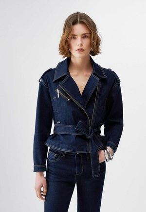 Donna con capelli corti e ondulati indossa una giacca di jeans blu scuro con vita a cintura, dettagli con cerniera, jeans in denim coordinati e bracciali d'argento al polso sinistro.